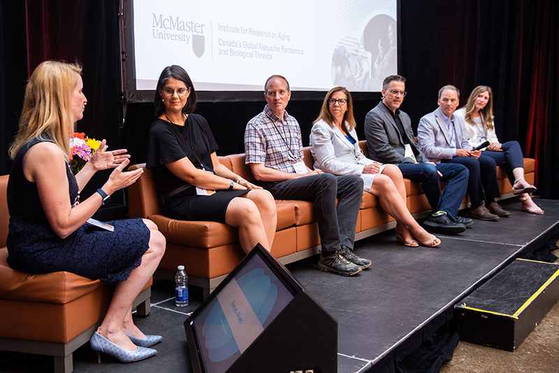 Sitting on couches on a stage, Rebecca Ganann addresses a panel of guests including Jennifer Walker, Arthur Sweetman, Doug Oliver, Laura Griffith, Jonathan Bean and Marla Beauchamp