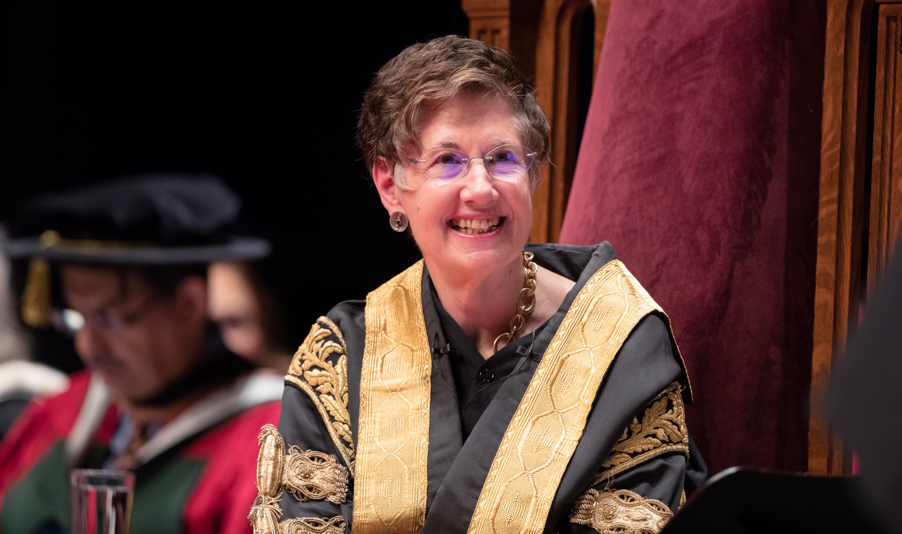 Person in the ornate gold and black academic robes of McMaster's Chancellor sits in a carved wooden chair with red upholstery, wearing glasses and a gold chain, with another robed figure blurred in the background.